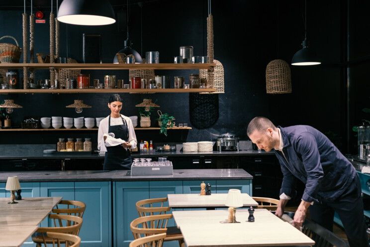 I et moderne køkkenmiljø med mørke vægge og træaccent arbejder to personer. En kvinde i en hvid skjorte står bag en køkkenø og forbereder ingredienser, mens en mand i en mørkeblå skjorte rengør et bord i forgrunden. Køkkenet er veludstyret med diverse køkkenredskaber og krukker på hylderne i baggrunden, der skaber en hyggelig og professionel atmosfære.
