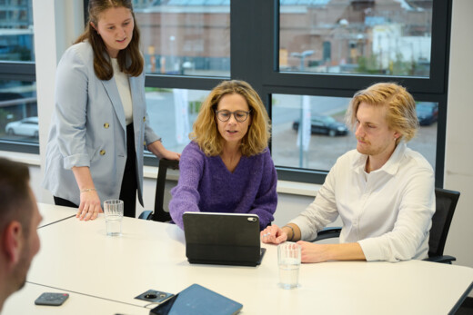 Three people looking at a tablet, two sitting down and one standing next to them.