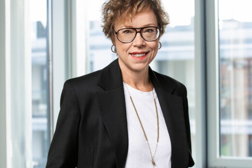 A woman wearing glasses and a black blazer, standing indoors by large windows with hands in pockets and a light-colored shirt.
