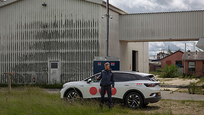 Securitas security guard stands with a mobile phone in his hand in front of his car on a company premises