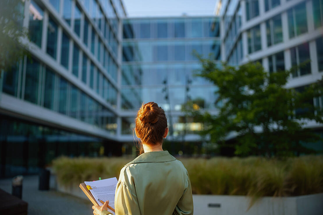 Woman holding a report looking to the office building surrounded by greens