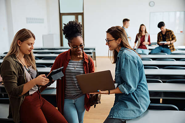 Grupo de estudiantes en un aula compartiendo información con una tableta y una computadora portátil, reflejando un ambiente educativo y colaborativo.