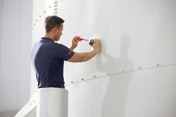 A Securitas technician installs a dome security camera onto a white industrial wall using a screwdriver.