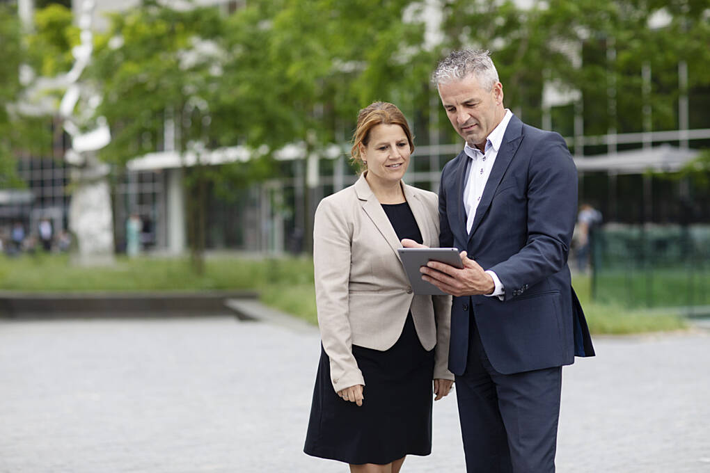 Két üzleti öltözetű szakember kültéri környezetben egy tablet képernyőjét tekinti át, irodai épület közelében, konzultációs helyzetben.