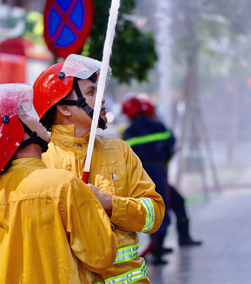 Two firefighters wearing yellow gear and red helmets direct a high-pressure stream of water upward, with a fire truck in the blurred background.