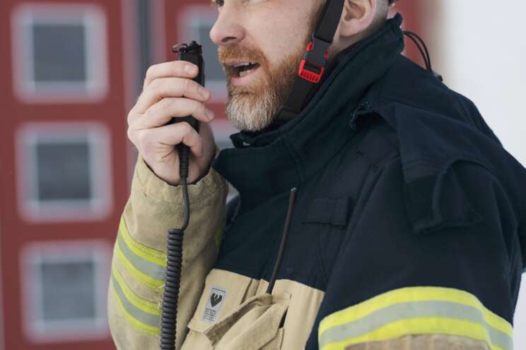 En brandmand fra Securitas med hjelm nummer 24 taler i en walkie-talkie, sandsynligvis koordinerer han en indsats under en nødsituation, med en bygning i baggrunden.