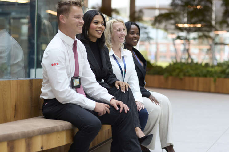 4 Mitarbeiter von Securitas sitzen auf einer Bank in einem Bürogebäude lächelnd nebeneinander