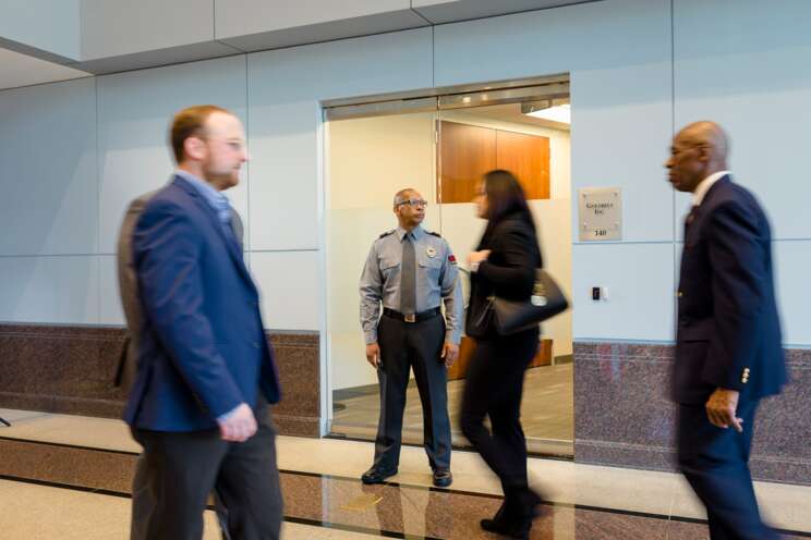Male security officer standing by an elevator observing people walking by.